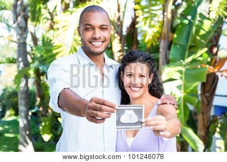 Portrait of happy husband with pregnant wife holding sonogram against white background