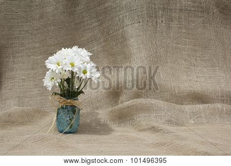 Bunch of daisies placed in a blue mason jar with a bow on a burlap background with space for type or other art