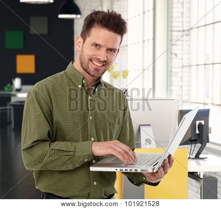 Portrait of happy smiling casual caucasian businessman working on laptop computer at creative office, standing, looking at camera.