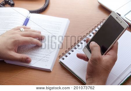 A businessman holds a smartphone in his hands. A phone a notebook a laptop and an open book lay beside him. Only hand in the frame