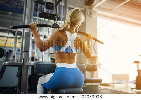 Beautiful sports young woman posing in fitness gym. Fitness girl in blue sportswear. Muscular blonde fitness woman doing exercises For Back On Machine. Sport - concept of healthy lifestyle.