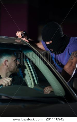 Closeup of dangerous criminal man with a gun in car parking. Auto theif threatening frightened driver, night time.