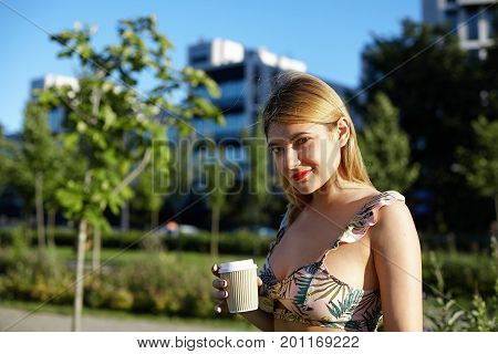Summer portrait of stylish young woman dressed in cropped top with floral pattern having walk in park on sunny day holding papercup with her favorite hot drink looking at camera with charming smile