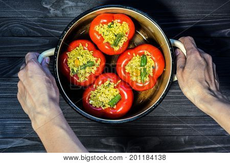 Chef holding a pan with four red stuffed peppers on dark rustic kitchen table background first person view. Cooking process top view