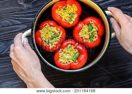 Chef holding a pan with four red stuffed peppers on dark rustic kitchen table background first person view. Cooking process top view