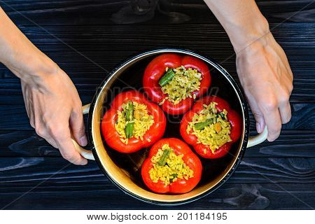 Chef holding a pan with four red stuffed peppers on dark rustic kitchen table background top view. Cooking process