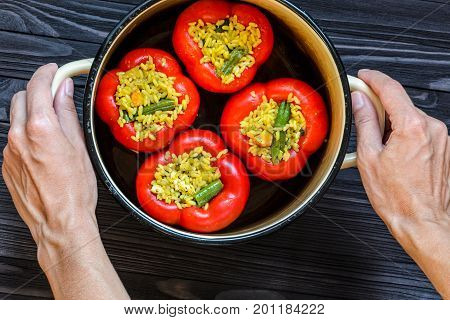 Chef holding a pan with four red stuffed peppers on dark rustic kitchen table background first person view. Cooking process top view