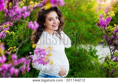 Pregnant woman walking in the spring park. With one hand she hugs belly and touches flowers with other, she is looking on the flowers and smell them