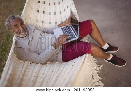 Portrait of smiling senior man using laptop while relaxing on hammock