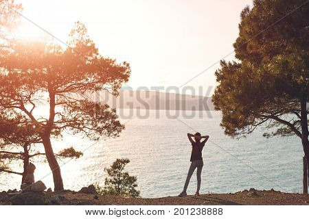 Back view of woman standing on the sea coast Between pines and Enjoying the sunset