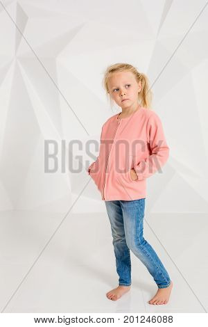Cute little girl in pink jacket and jeans, hands in pockets on white background. Little brunette posing in studio