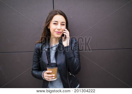 Beautiful Student Girl Leaning On Wall, Talking On Smart Phone And Holding Takeaway Coffee In Paperc