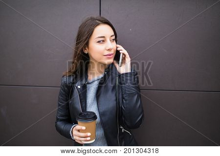 Beautiful Student Girl Leaning On Wall, Talking On Smart Phone And Holding Takeaway Coffee In Paperc