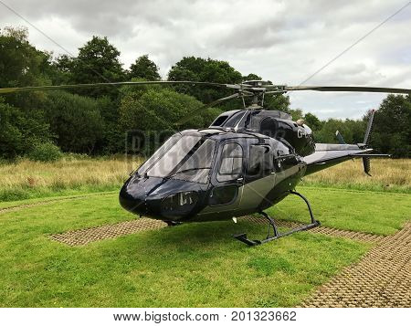 Front side view of a black squirrel helicopter that landed on the H helicopter landing spot. With grass and trees in the background on a cloudy day.