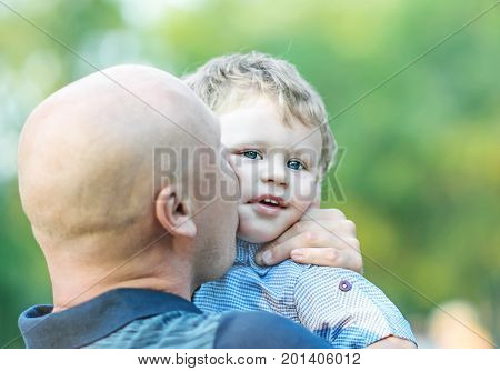 A happy father embracing and kissing his adorable small child outdoor at summer park. Blurred background