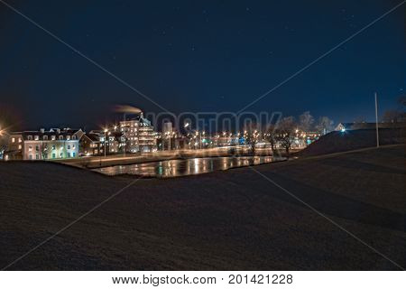 Night cityscape with a lawn in the foreground, lake in the middleground, and a city in the background