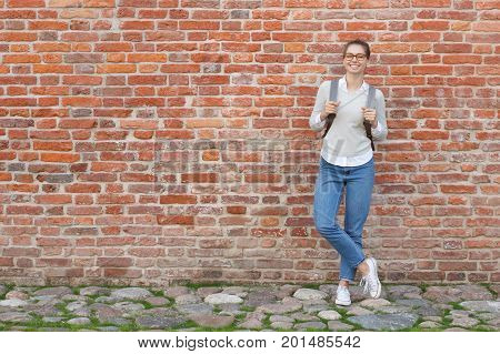Full-height Portrait Of Young European Female Standing Against Red Brick Wall, Leaning To It With Ba