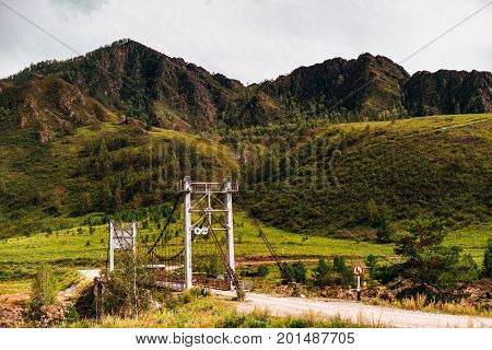 Rope bridge over the most narrow part of Katun river with mountain-ridge and green meadows in background and dirt road in foreground Altai mountains Kuyus district Russia