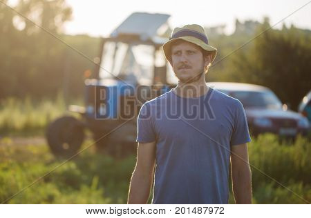 Portrait of young farmer in hat on the field in sunset light with the tractor on the background at organic eco farm.
