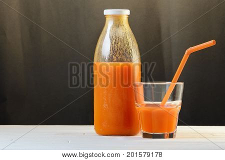 A bottle of carrot juice and a glass of carrot juice with a straw on white wood table on black background. Still life of a healthy eating concept
