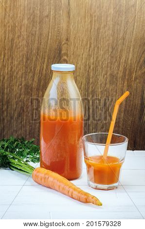 A bottle of carrot juice and a glass of carrot juice with a straw on white wood table against brown wood wall background. Still life of a healthy eating concept