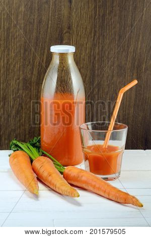 A bottle of carrot juice and a glass of carrot juice with a straw on white wood table against brown wood wall background. Still life of a healthy eating concept