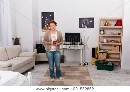 View of mother in the room of her teenager son. Mid aged foxy woman standing in light room holding red cup in her hands.