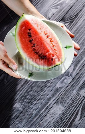 Slice of watermelon on white plate in woman hands ready for eating top view with blurry black wood table background