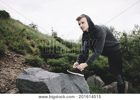 Well built runner is taking a short break. He has put his right leg on the top of the stone and tying his sneakers. The guy is ready to continue jogging as soon as possible.