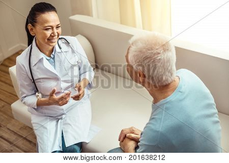 Feeling comfortable. Positive nice friendly nurse sitting on the sofa and talking to her patient while asking him about his health