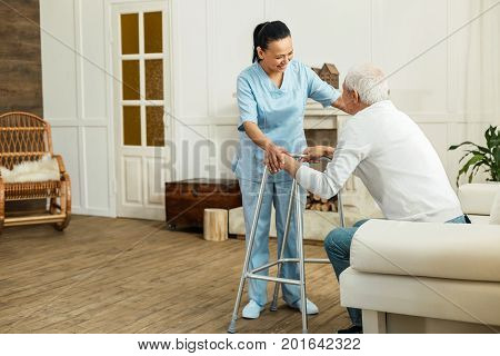 I will help you. Joyful delighted professional caregiver smiling to her patient and helping him to get up while doing her job