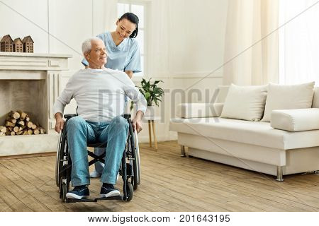 Time for a walk. Cheerful positive female caregiver standing behind her patient and moving a wheelchair while taking him for a walk