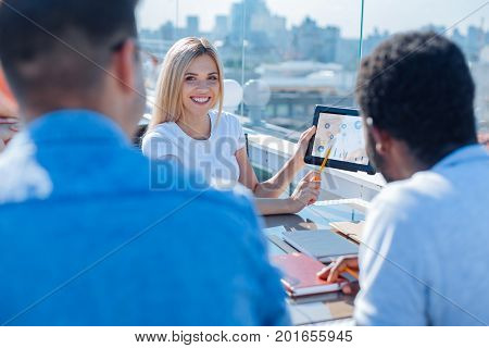 Here is the result. Selective focus on a beaming young woman holding a tablet and pointing at a screen of a gadget while working together with her coworkers outdoors.