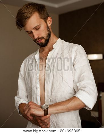 Young man in white unbuttoned shirt dresses fasten watchband.