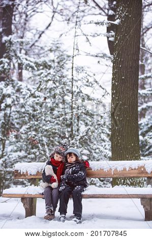 Two Children, Boy Brothers, Sitting On A Bench In Park, Wintertime While Snowing
