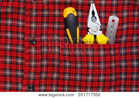 Used work hand tools - pliers screwdriver and ruler - in checkered shirt pocket. Work outfit. Horizontal close-up capture.