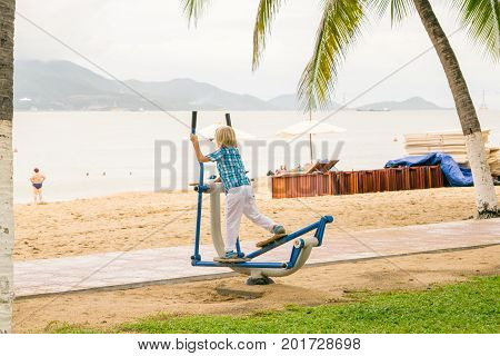 A blond boy does excersices at training device in a park on a beach, Nha Trang, Vietnam