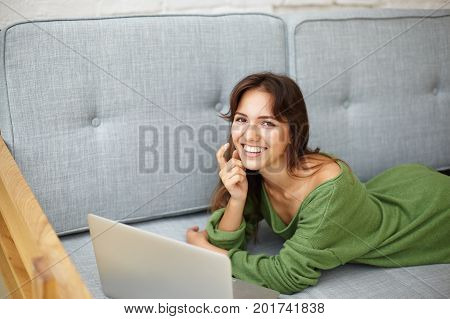 Always connected. Indoor picture of student girl spending leisure time after lectures at college lying on couch in living room surfing internet on her web enabled generic laptop computer pc