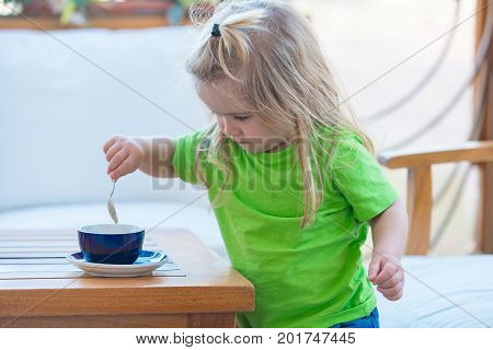 Child boy with blond long hair mixing sugar with teaspoon. Little kid boy making tea. Boy child mixing sugar in blue cup. Healthy breakfast for child. Childhood and baby care concept.