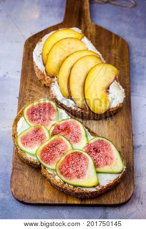 Toasted sandwiches with cream cheese fresh ripe green and red figs and peaches. Drizzled with honey. Wholegrain rye bran bread on wood cutting board grey concrete background
