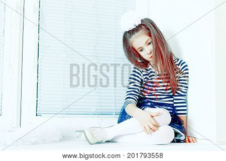Little girl in blue striped dress and a white bow on her head.She sits at the window with sad eyes something lost in thought.Creative toning of a photograph.