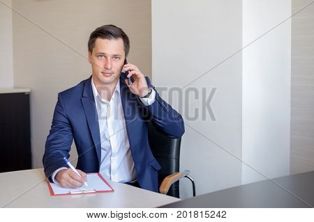 serious business man in office making cell phone call and taking notes.