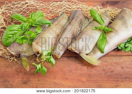 Several uncooked carcasses of the notothenia fish without of a heads and tails and twigs of basil and parsley closeup on the fishing net on a dark wooden surface