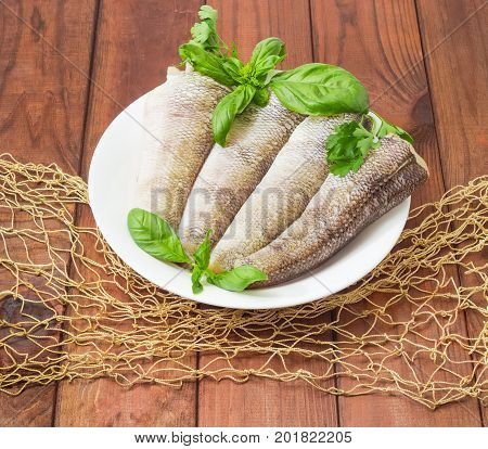Carcasses of the notothenia fish without of a heads and tails and with peeled scales and prepared for cooking basil and parsley twigs on the white dish on a dark wooden surface with fishing net