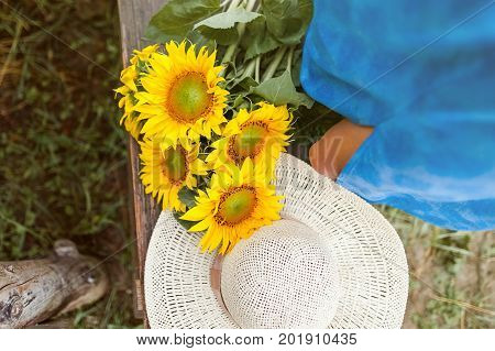 Top view of a sun flower bouquet and a straw hat on a wood banch and a woman standing next to it, wearing a blue dress.