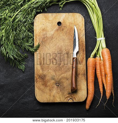 A bunch of fresh juicy carrots with a tops on a dark background with a vintage cutting board and a knife for cooking dinner. Top View