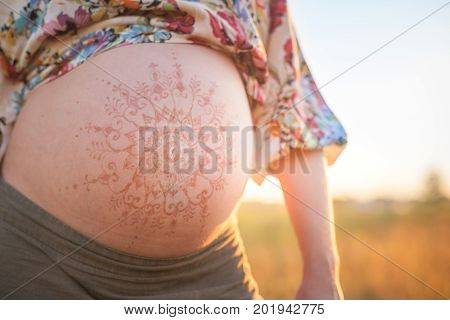 A beautiful pregnant woman on the beach touching her mehandi ornament belly with love and care at sunset lights summer evening