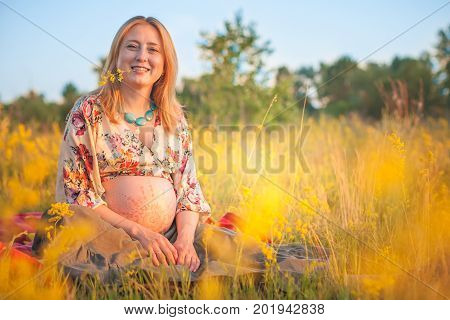 9 months pregnant woman with mehandi pattern ornament belly sitting in yellow grass and smiling on sunset lights. Waiting for baby. Pregnancy concept