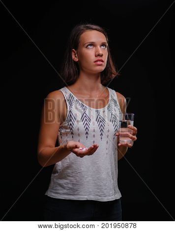 Exhausted young woman looking up in pain on the black background. A girl with short dark hair, in a light shirt holding many pills and a glass of cold water. Medictional treatment concept.