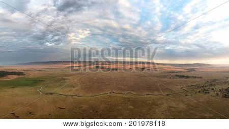 Panorama of the open plains of the Masai Mara at daybreak, with the Mara river and the hills of the Oldoinyio escarpment. Aerial view from a balloon. Hot air balloons can be seen in the distance.